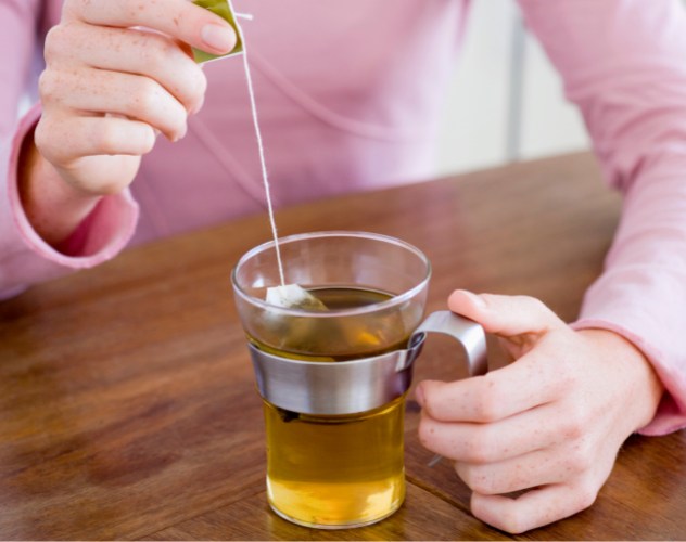 person in a pink long sleeve top stirring a clear mug of herbal tea on wooden table